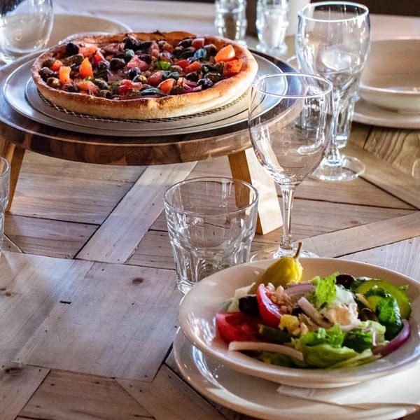 A deep-dish pizza with tomatoes and olives on a stand, next to a fresh salad and glasses.