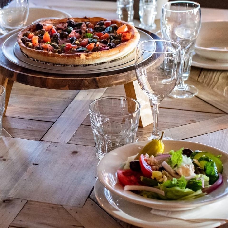 A deep-dish pizza with tomatoes and olives on a stand, next to a fresh salad and glasses.