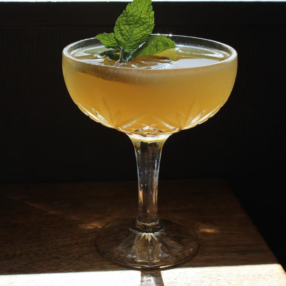 A golden cocktail in a cut-glass coupe, garnished with fresh mint and a citrus slice.
