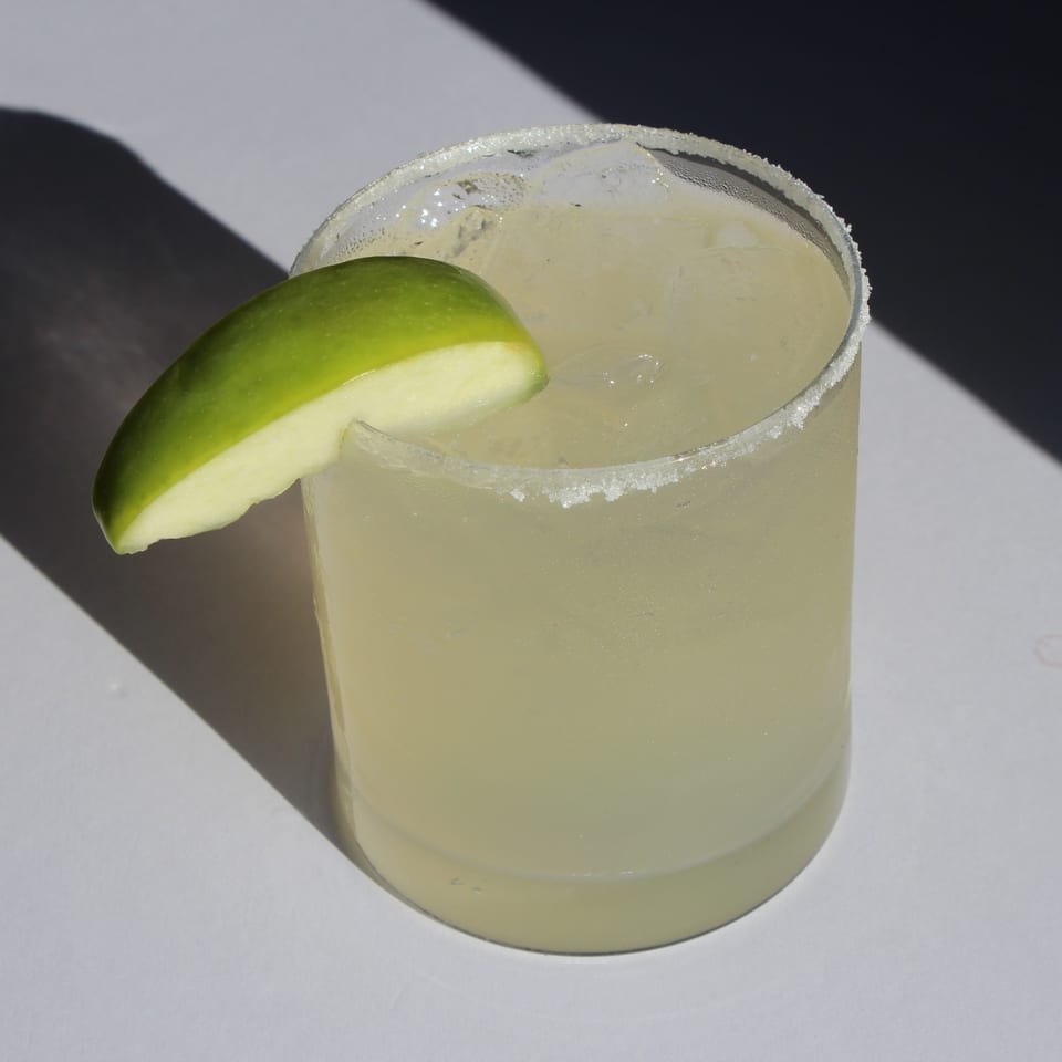 A margarita with a salted rim and a green apple slice, casting a shadow on a white surface.