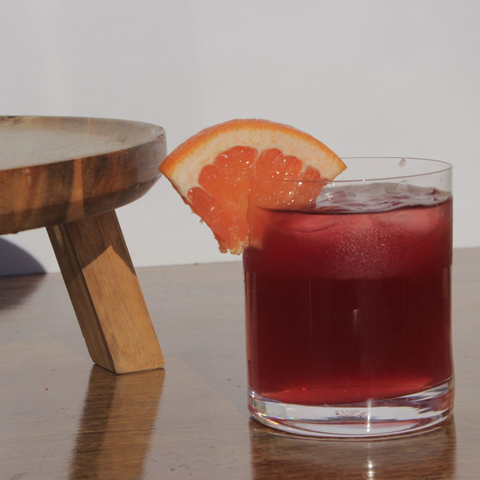 A glass of red sangria with ice and a grapefruit slice, next to a wooden stool.