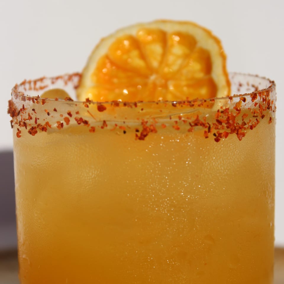 Close-up of a vibrant orange cocktail with a chili-salted rim and an orange slice garnish.