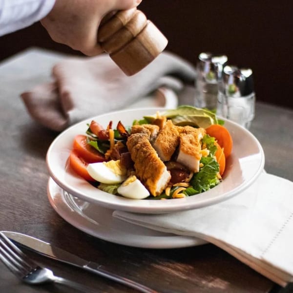 A hand seasons a fresh salad with crispy chicken, tomatoes, avocado, and hard-boiled eggs.