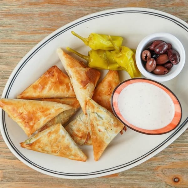 Golden-brown triangular pastries, green peppers, black olives, and a white dipping sauce on a white plate.